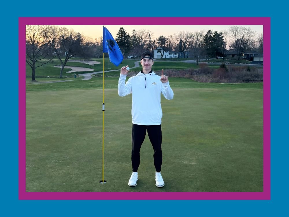 Ryan Kardell holding golf ball and holding up one finger to signify scoring a hole-in-one